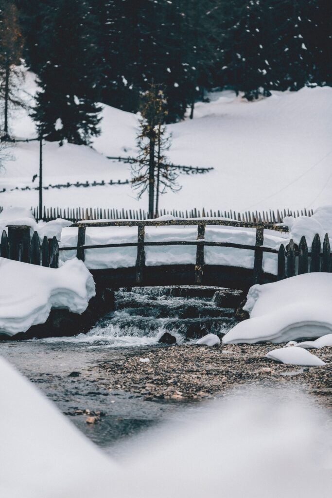 Rustic wooden bridge over creek in serene snowy forest with bare trees and soft snowfall winter scene Wallpaper