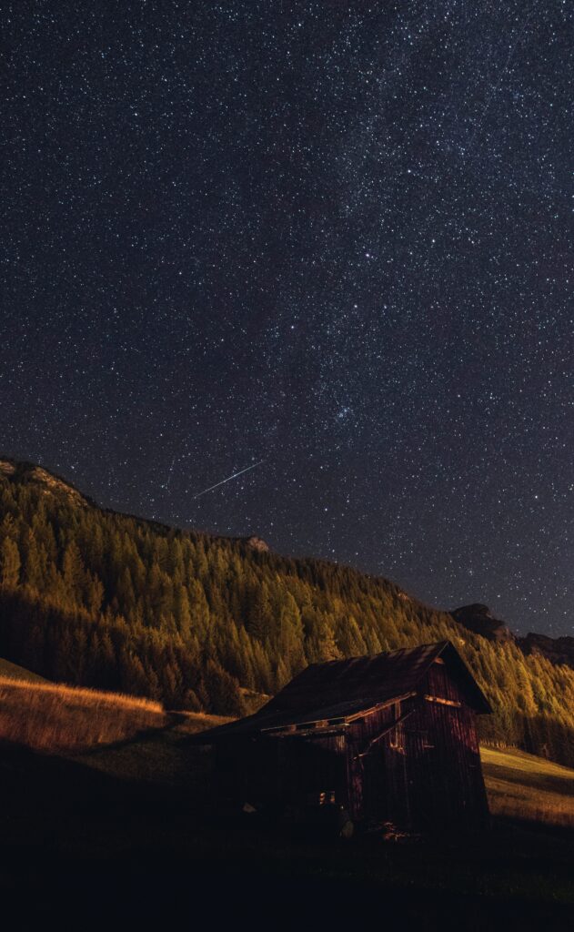 Rustic wooden cabin by dense forest under starry night sky with shooting star peaceful wallpaper
