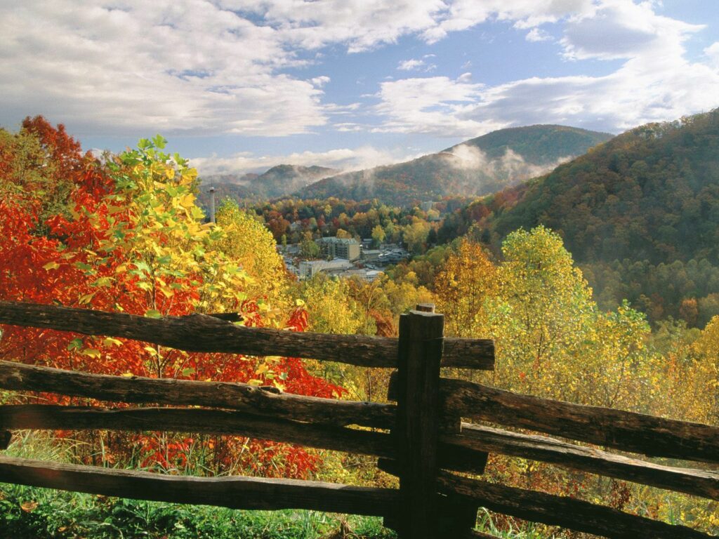 Rustic log fence with vibrant autumn trees rolling hills and village under soft sunlight background Wallpaper