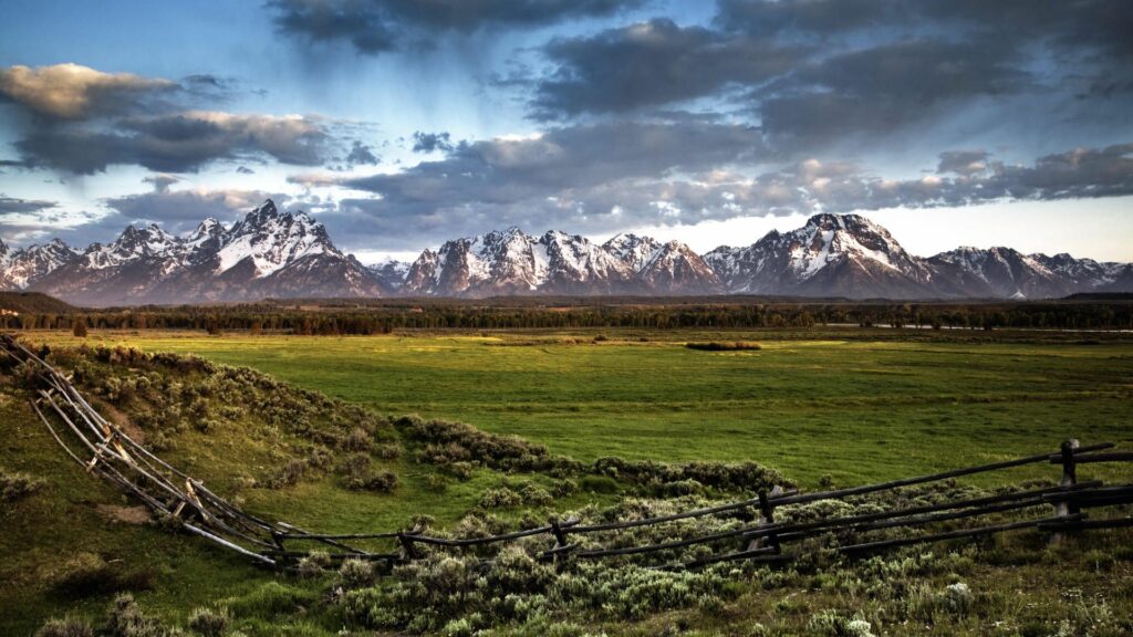 Rustic wooden fence across green field with snow-capped mountains and textured cloudy sky Wallpaper