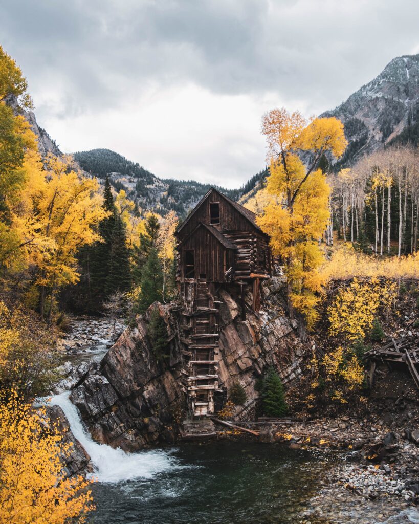 Rustic wooden hut on rocky hill by winding river with autumn trees and distant mountains view Wallpaper