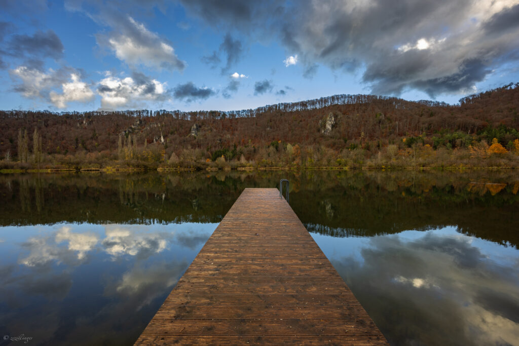 Rustic wooden pier over calm autumn lake reflecting forest and dramatic cloudy sky wallpaper background