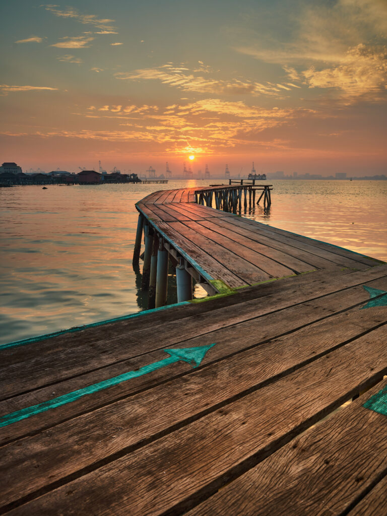 Rustic wooden pier over calm water at sunset with vibrant port cranes and golden reflections Wallpaper