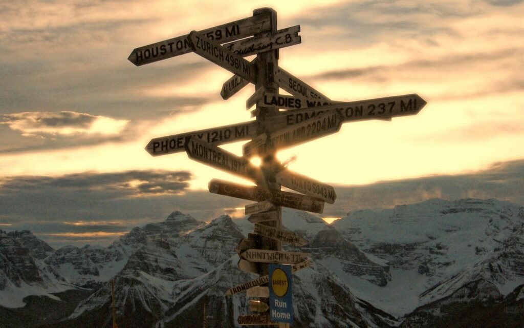 Rustic wooden signpost with city names against snow-capped mountains and glowing sunset sky wallpaper