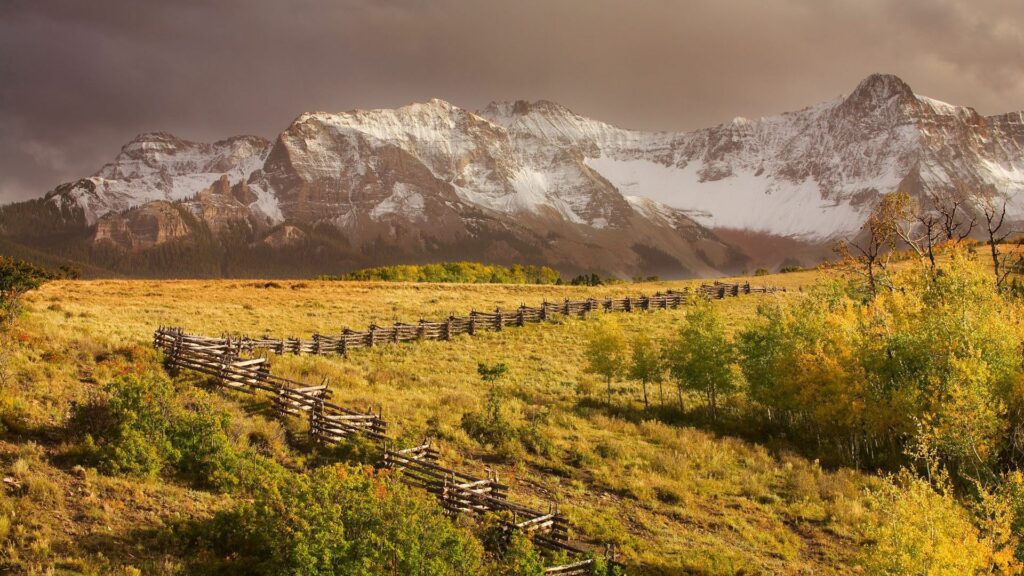 Rustic zigzag fence in golden autumn fields with vibrant trees and snow-capped mountains background Wallpaper