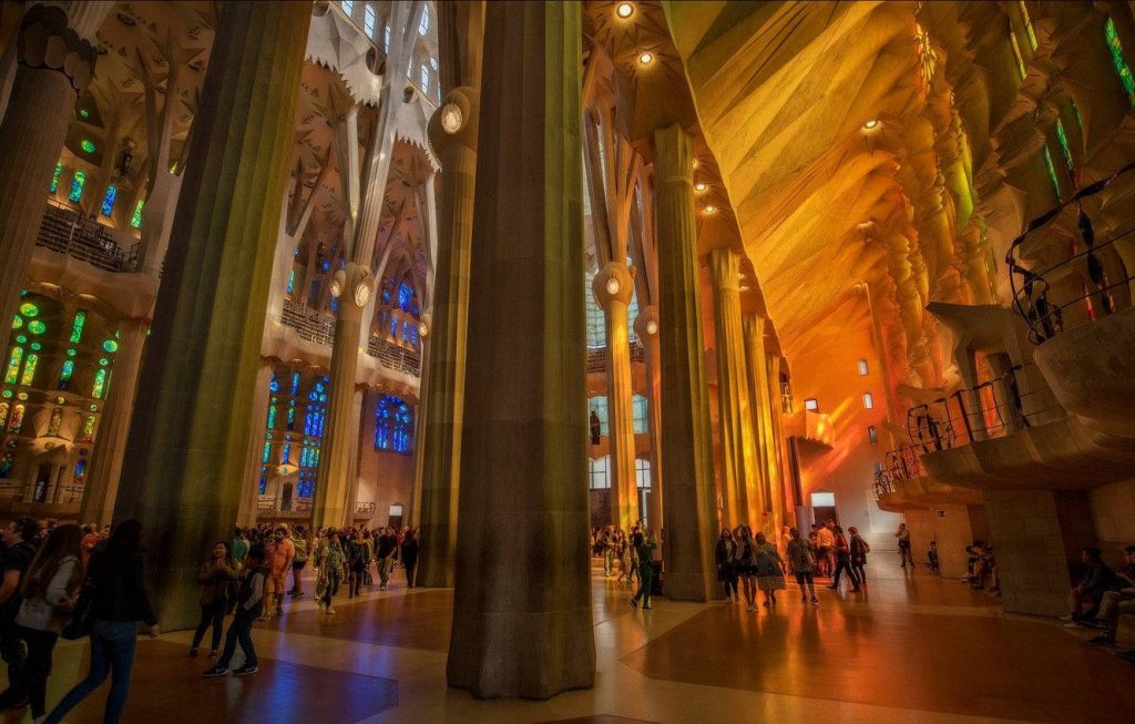 Sagrada Familia interior with vibrant stained glass light on tree-like stone columns and tourists Wallpaper
