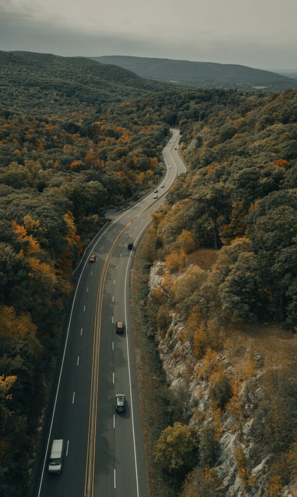 Winding highway through dense orange and green autumn forest with cars on scenic fall drive Wallpaper