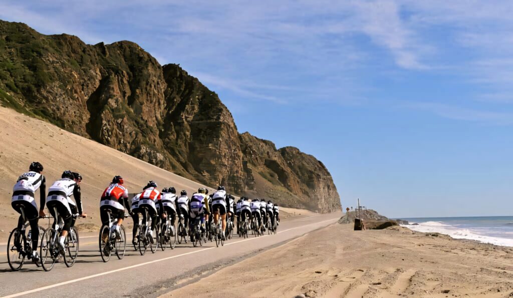 Cyclists riding scenic coastal road with cliffs ocean bright blue sky vibrant landscape wallpaper