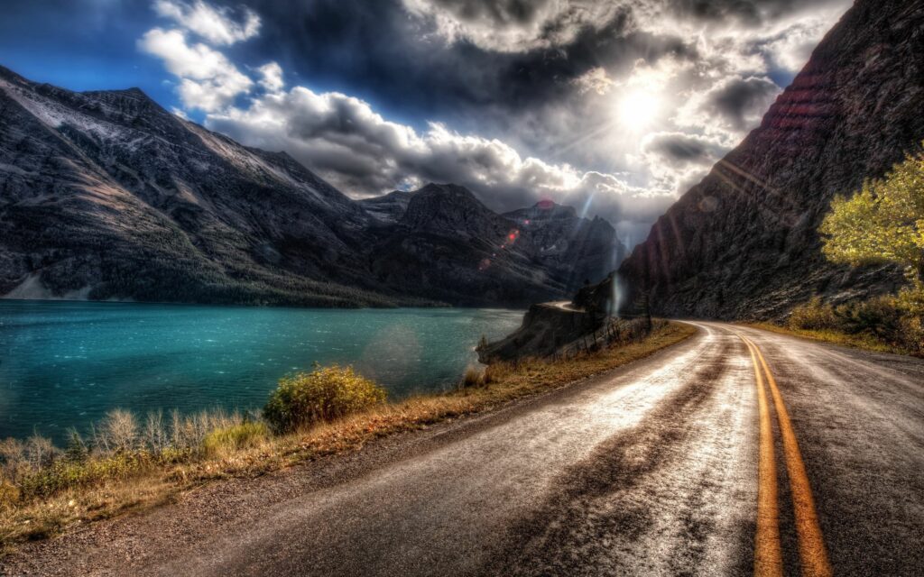 Scenic asphalt road with double yellow lines winding along turquoise river and rugged coast under dramatic sunlight Wallpaper