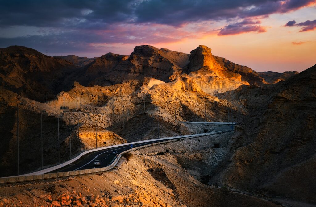 Winding mountain road with cars under golden sunset sky and rugged rocky cliffs landscape Wallpaper