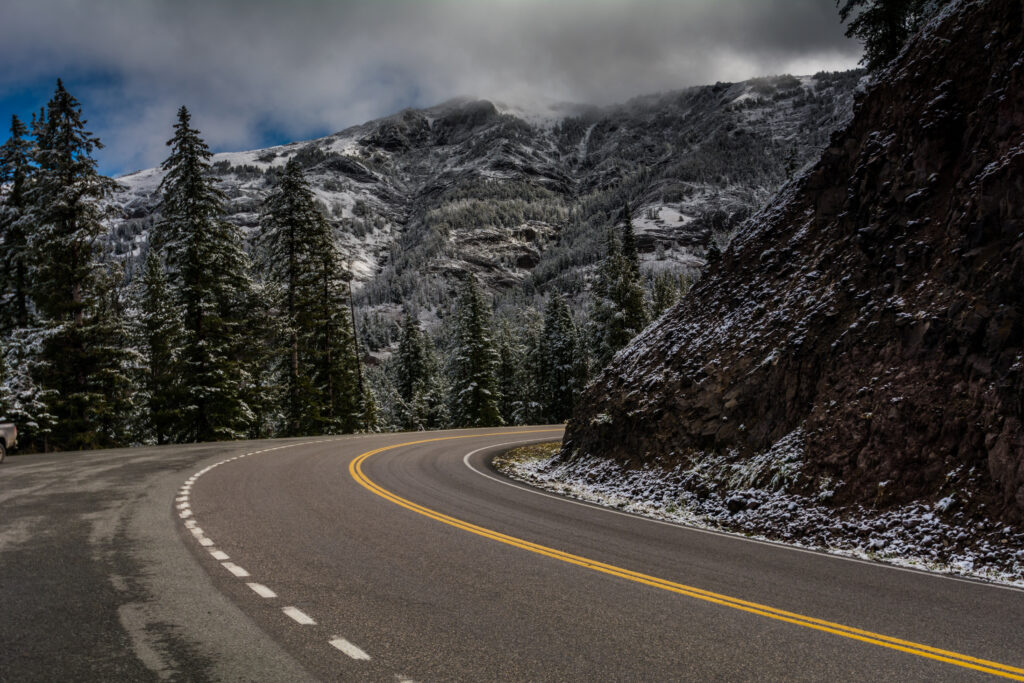 Mountain road winding through snowy trees and rocky cliffs under dramatic cloudy sky wilderness view Wallpaper
