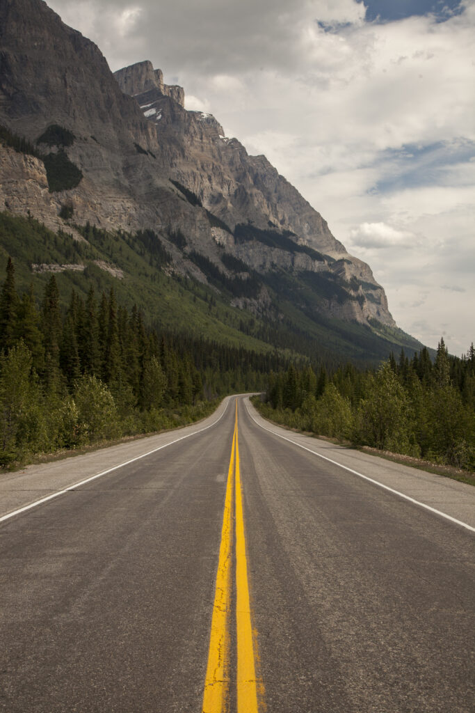 Straight road through green trees toward rugged mountain cliffs under a partly cloudy sky wallpaper