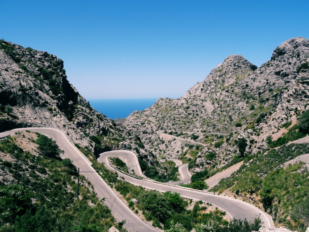 Winding mountain road through cliffs and green grass under blue sky with sea horizon view Wallpaper