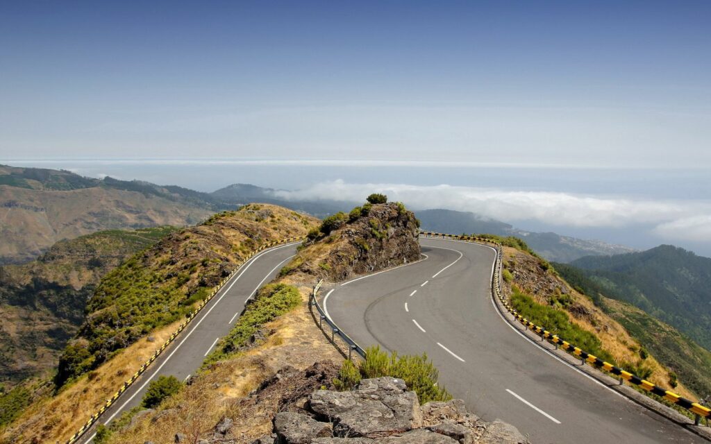 Winding mountain road with yellow black barriers overlooking hills and clouds under blue sky Wallpaper