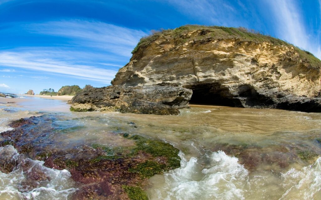 Scenic river flowing past mossy stones to rugged rock cave under vibrant blue sky with clouds Wallpaper