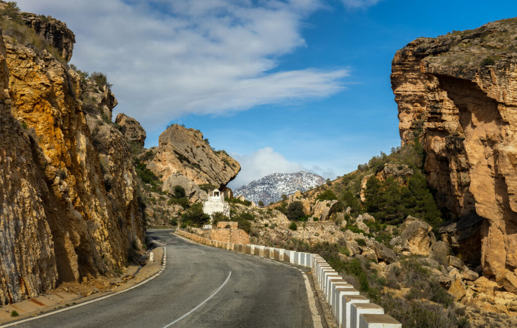 Scenic road between golden rock formations with chapel and snow-capped mountains under blue sky Wallpaper