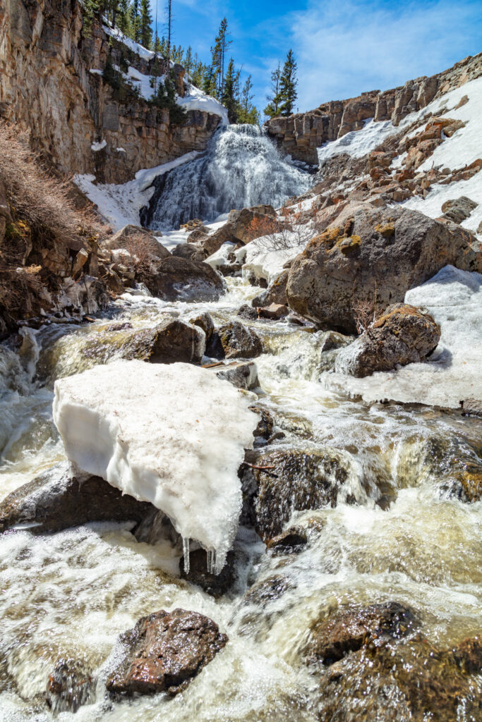 Scenic waterfall over rocky stones with melting snow and evergreen trees under blue sky Wallpaper