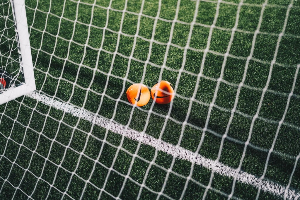 Two vibrant orange Nike soccer balls on green turf behind a white goal net stadium wallpaper