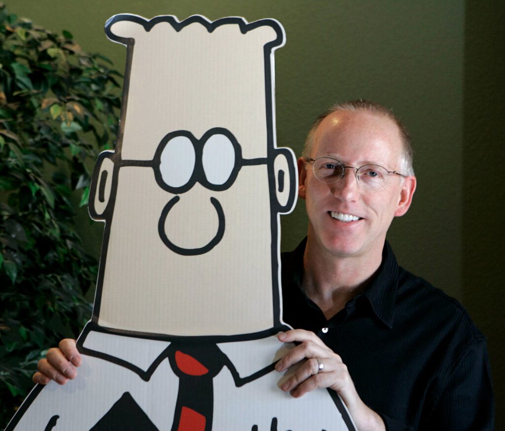 Scott Adams standing next to large Dilbert cutout in warm creative setting with natural colors Wallpaper