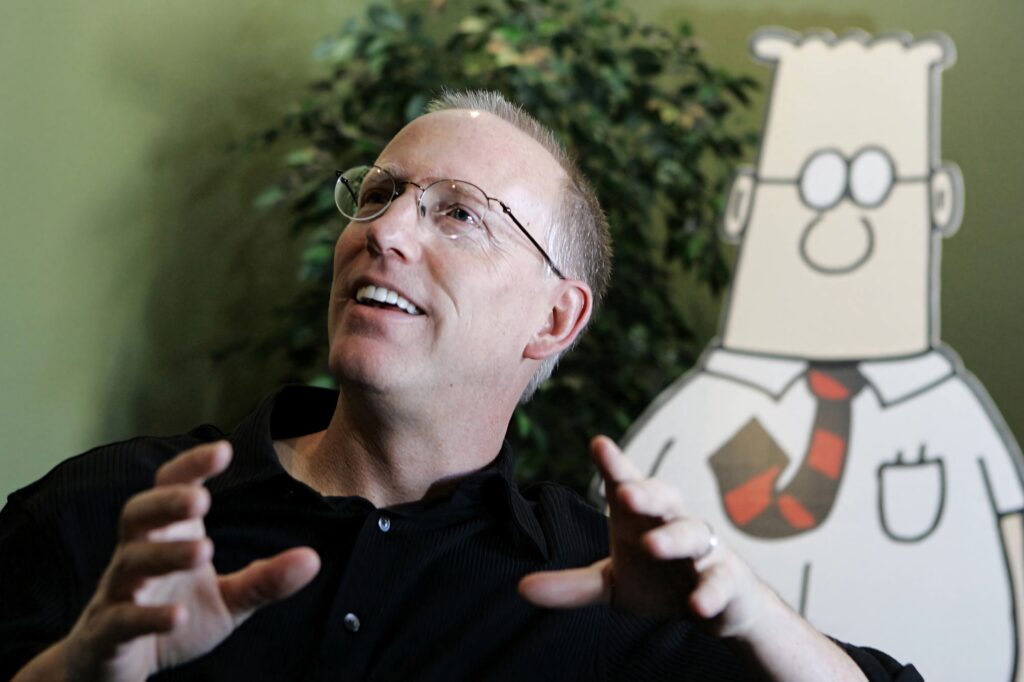 Scott Adams smiling beside Dilbert cutout in creative workspace with natural light and greenery Wallpaper