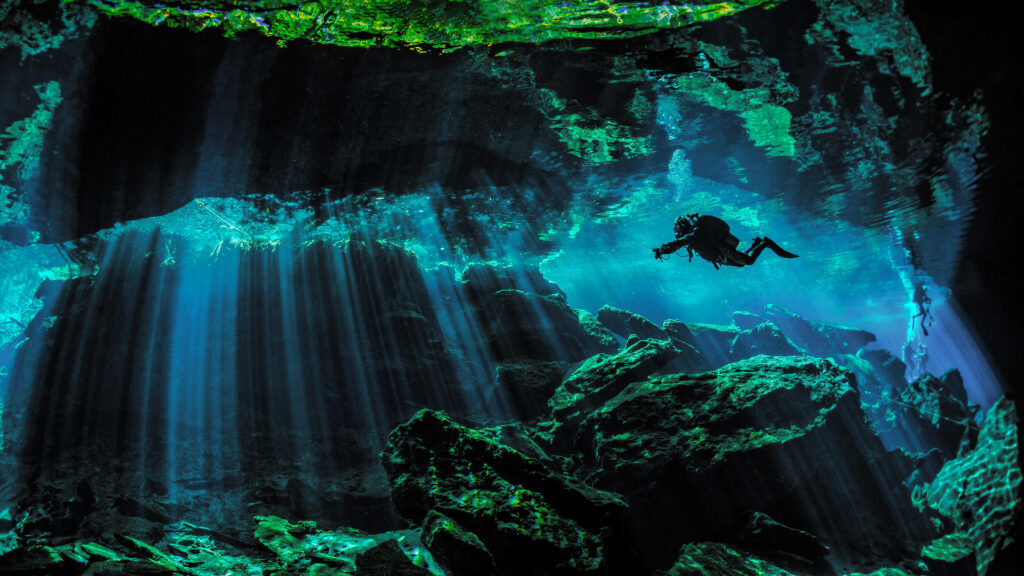 Scuba diver exploring illuminated underwater cave with vibrant green rocks and blue water wallpaper