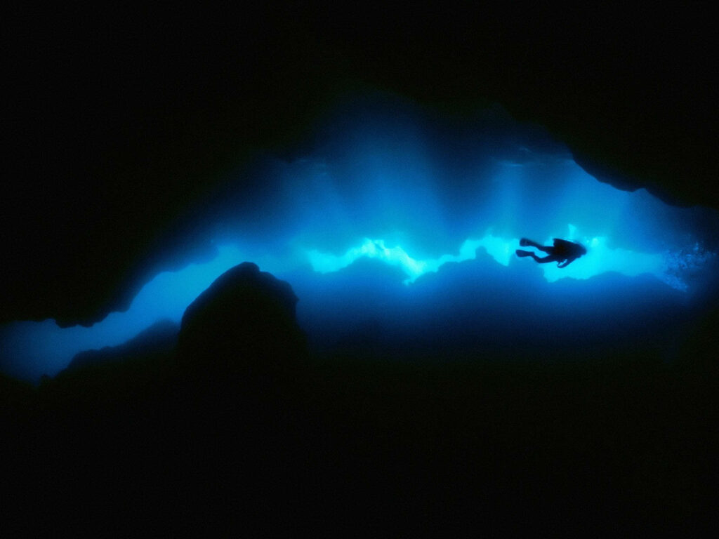 Scuba diver exploring illuminated underwater cave with blue light and dark silhouettes background Wallpaper