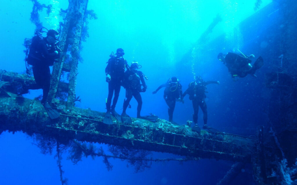 Scuba divers explore vibrant sunken shipwreck with sunlight beams and marine life underwater Wallpaper