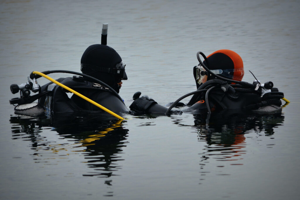 Two scuba divers in black wetsuits float on calm water discussing teamwork in underwater adventure Wallpaper