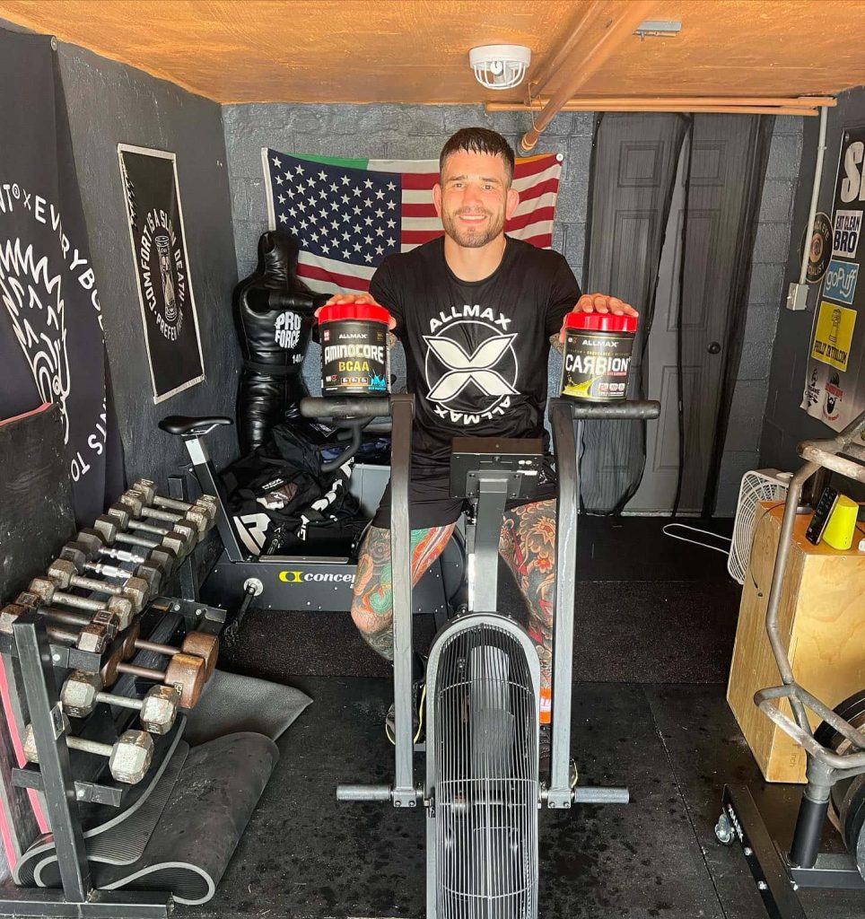 UFC fighter Sean Brady on stationary bike with Allmax supplements in rugged garage gym Wallpaper
