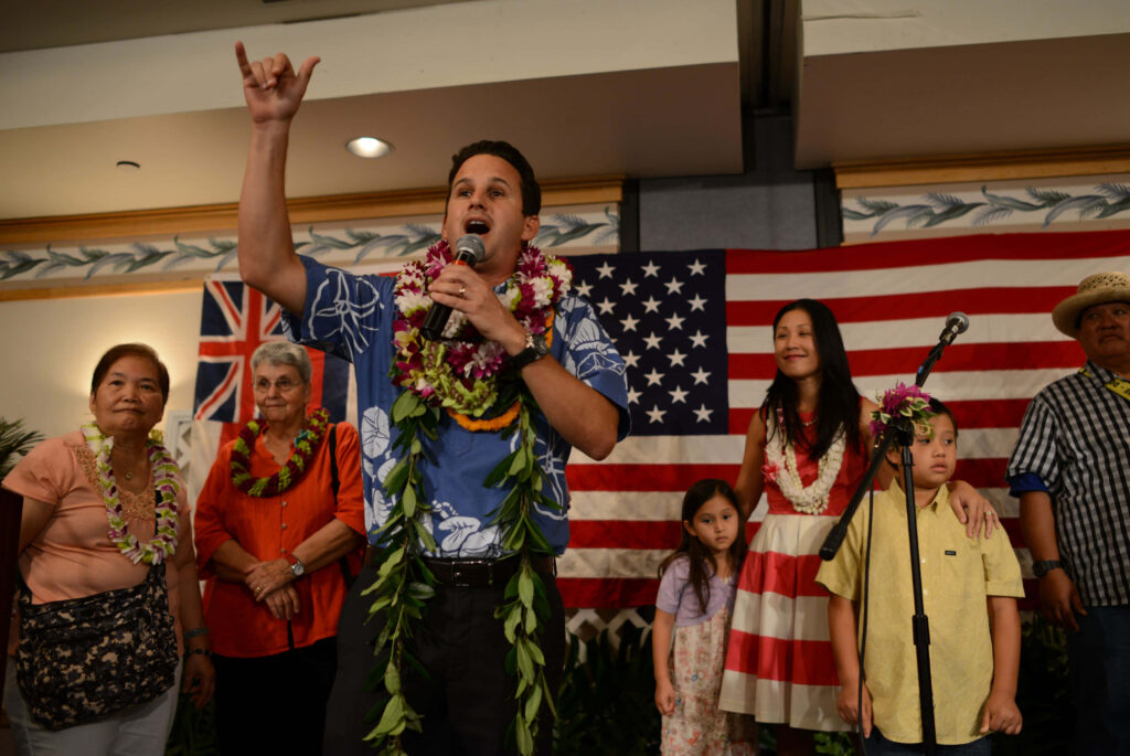 Senator Brian Schatz speaking with diverse supporters in Hawaiian leis against US and Hawaii flags Wallpaper