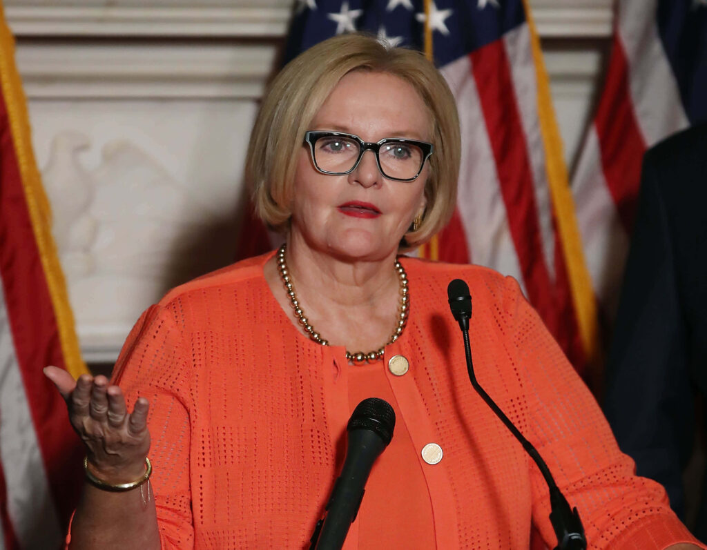 Senator Claire McCaskill in peach blazer stands confidently before American flags with poised expression Wallpaper