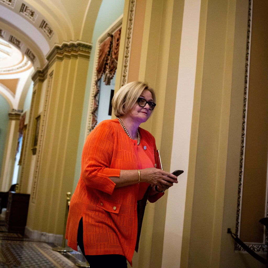 Senator Claire McCaskill walking Capitol Hill halls in orange jacket holding phone under warm light Wallpaper
