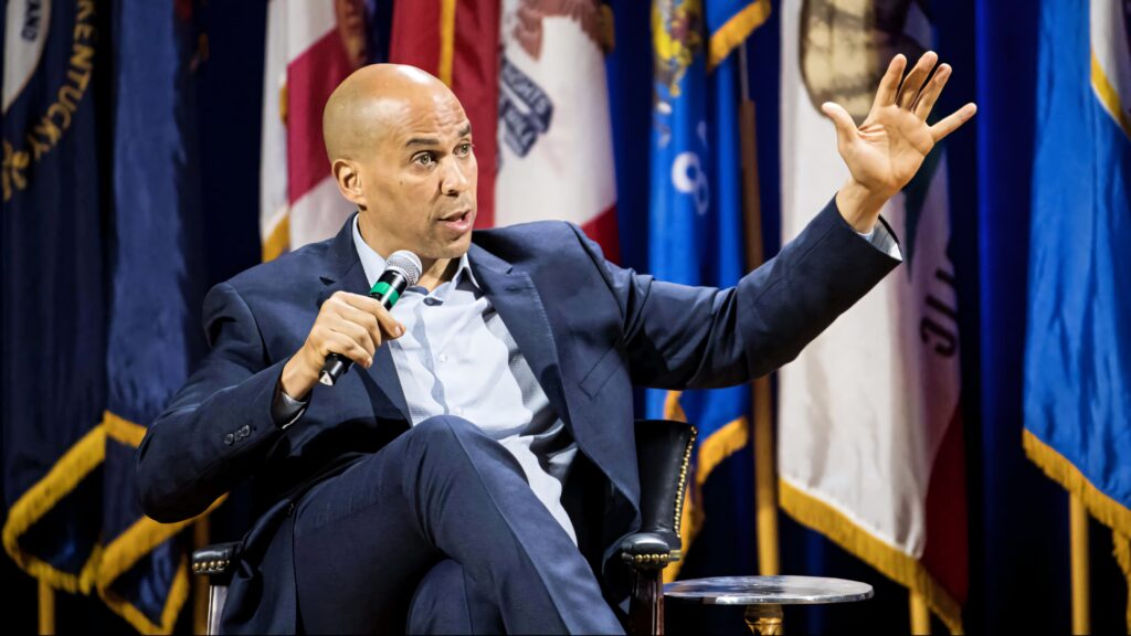 Senator Cory Booker speaking on TV set in dark suit with microphone American flags background Wallpaper
