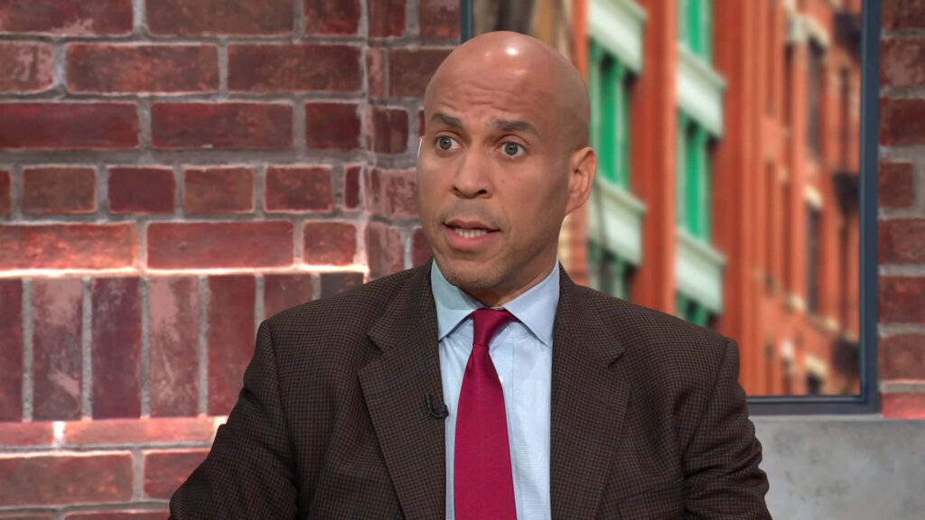 Senator Cory Booker speaking passionately in dark suit blue shirt red tie against brick wall background Wallpaper