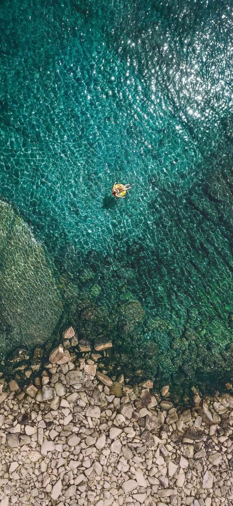 Aerial view of person on yellow float in clear turquoise water by rocky coast summer wallpaper background