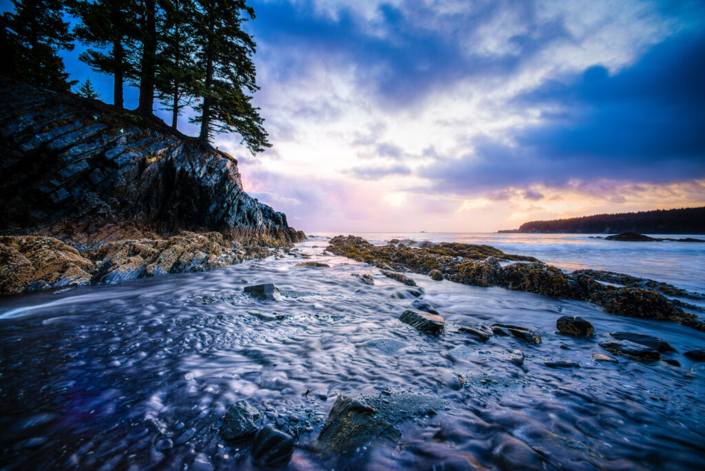 Serene Alaskan stream flowing through stones with trees cliffs and vibrant sunset sky wallpaper