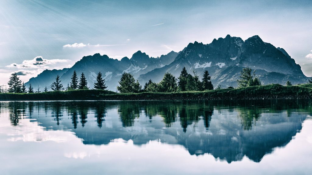 Crystal-clear mountain lake reflects snow-capped peaks and lush trees in serene alpine landscape Wallpaper