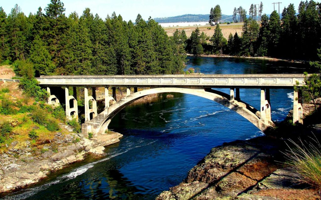 Classic arched stone bridge over clear river surrounded by lush green forest in bright sunlight Wallpaper