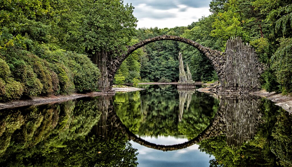 Arched stone bridge over calm river with lush green trees reflected for serene wallpaper background