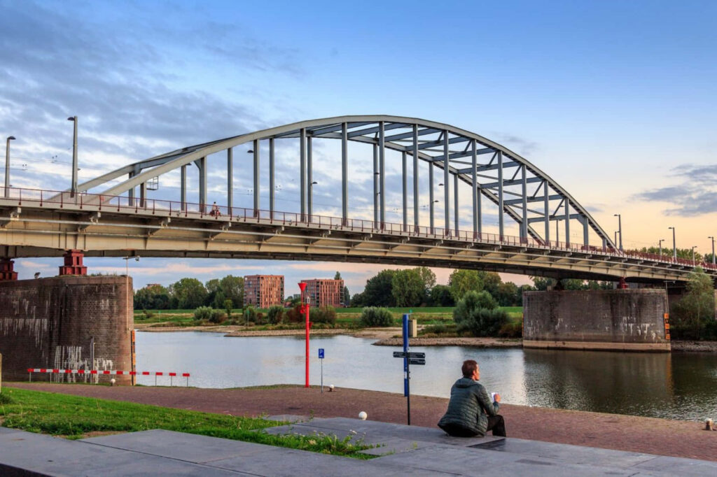 Serene Arnhem Bridge at dusk with evening light reflections and person relaxing by riverside Wallpaper