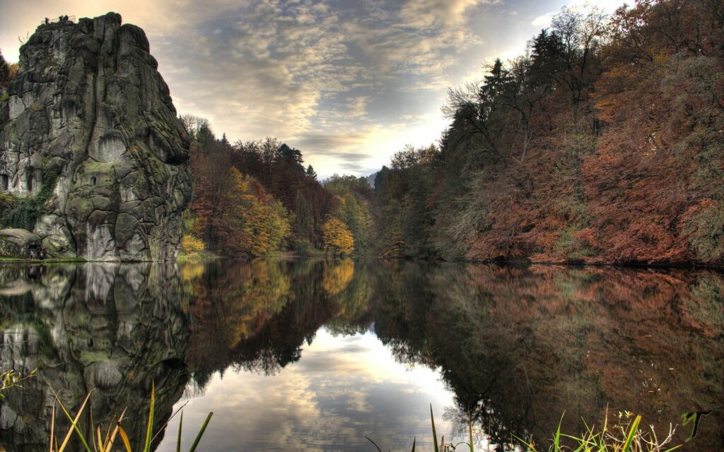 Gray rocky cliffs beside a calm lake reflecting autumn trees and a cloudy sky landscape wallpaper