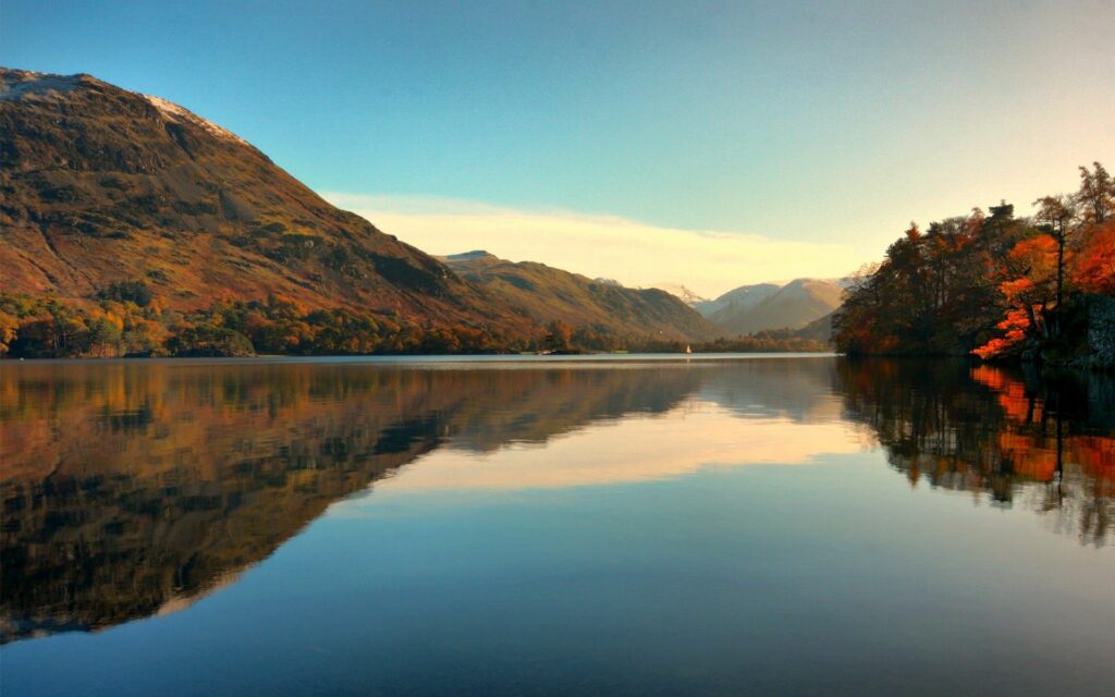 Serene autumn lakeside with calm water reflecting mountains and vibrant orange green trees Wallpaper