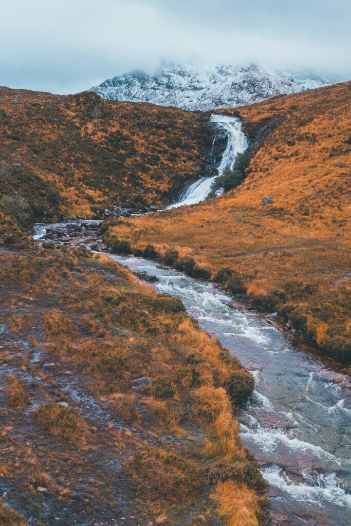 Crystal-clear stream winding through golden hills with waterfall and snowy mountains autumn wallpaper