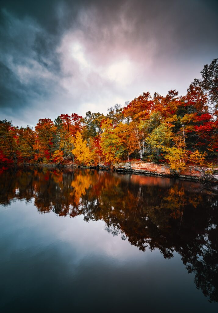 Serene autumn landscape with vibrant red orange gold trees reflected on calm lake under cloudy sky Wallpaper