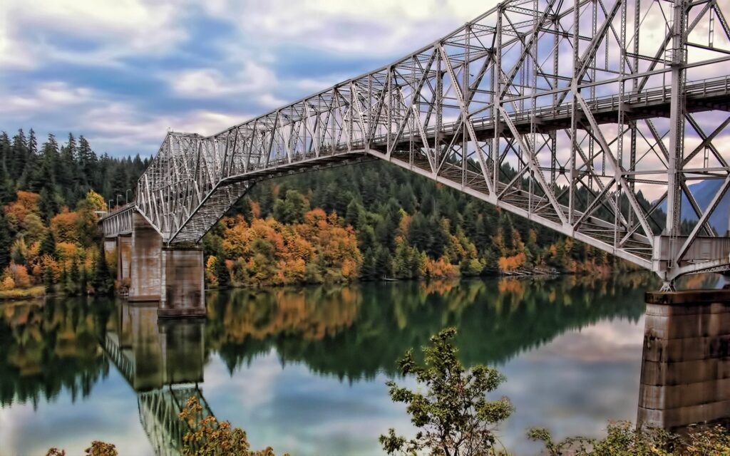 Metal bridge over calm river reflecting autumn foliage and green trees under cloudy sky wallpaper