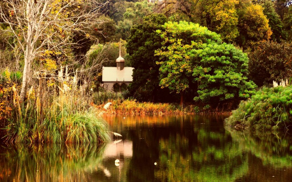 Serene autumn pond reflecting colorful trees with quaint chapel and lush green landscape wallpaper