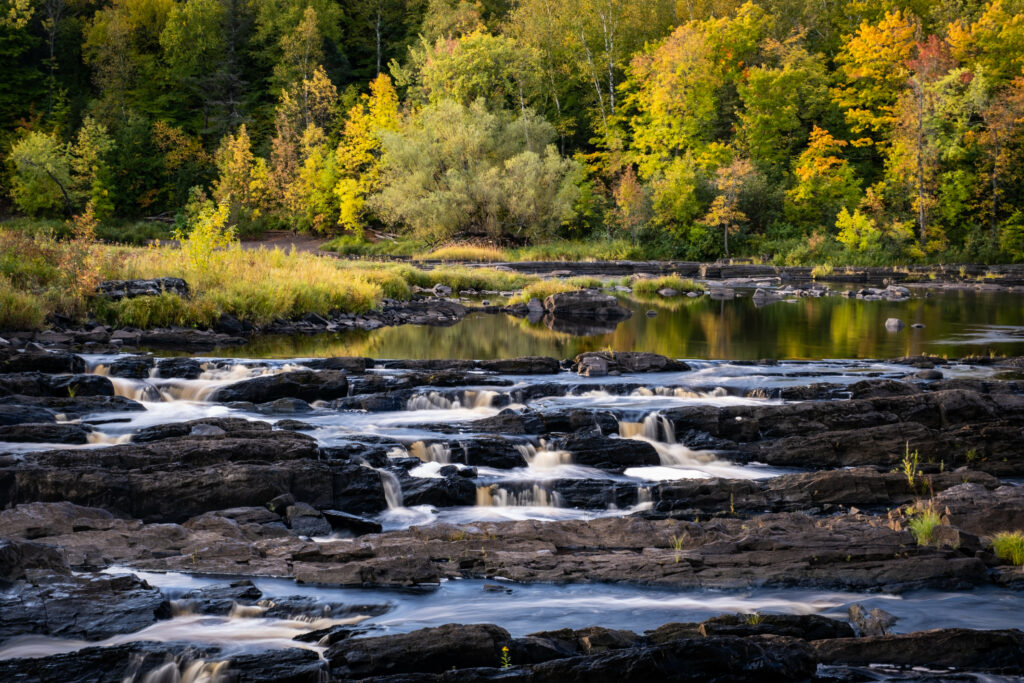Serene river flowing over dark rocks in lush forest with vibrant autumn colors and golden leaves Wallpaper