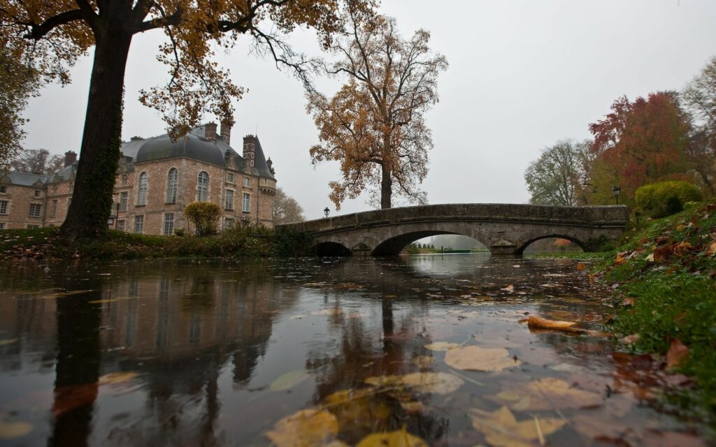 Serene riverside stone bridge reflecting autumn leaves and historic mansion under overcast sky Wallpaper