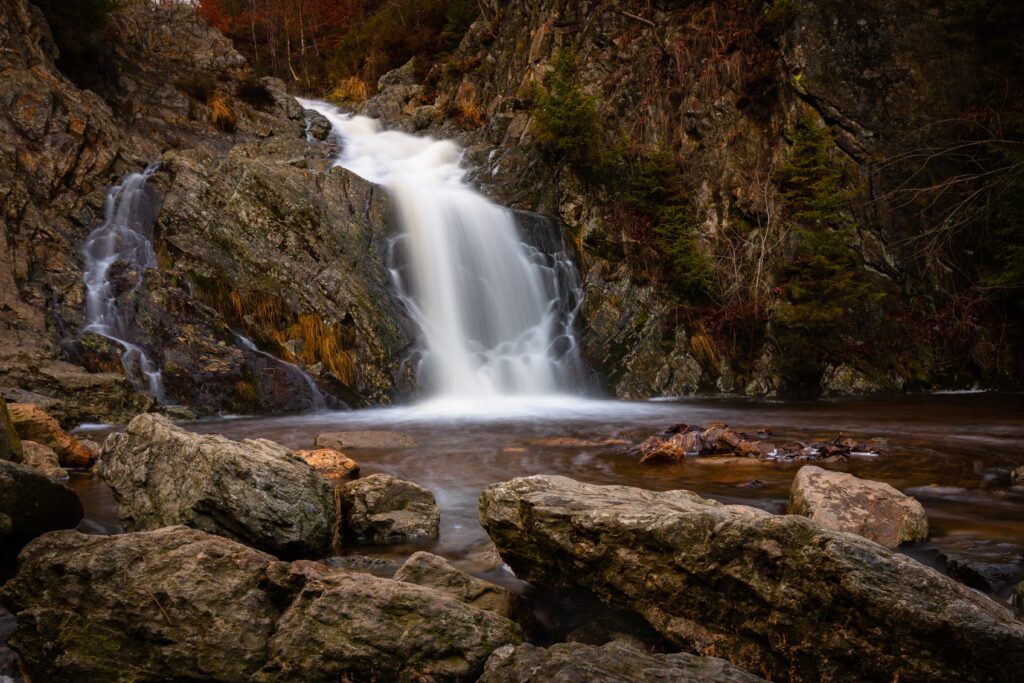 Serene waterfall over rugged grey stones flowing into calm pool with rich brown autumn hues wallpaper