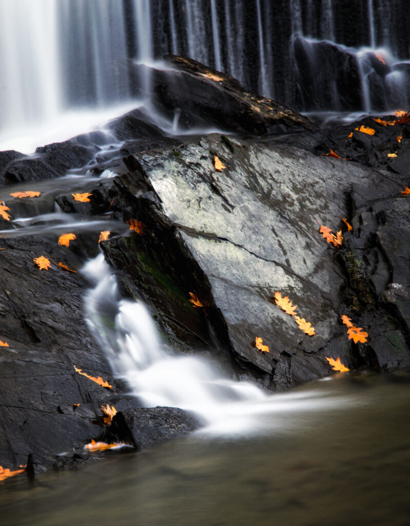 Serene autumn waterfall flowing over dark rocks with vibrant orange leaves and misty splashes Wallpaper